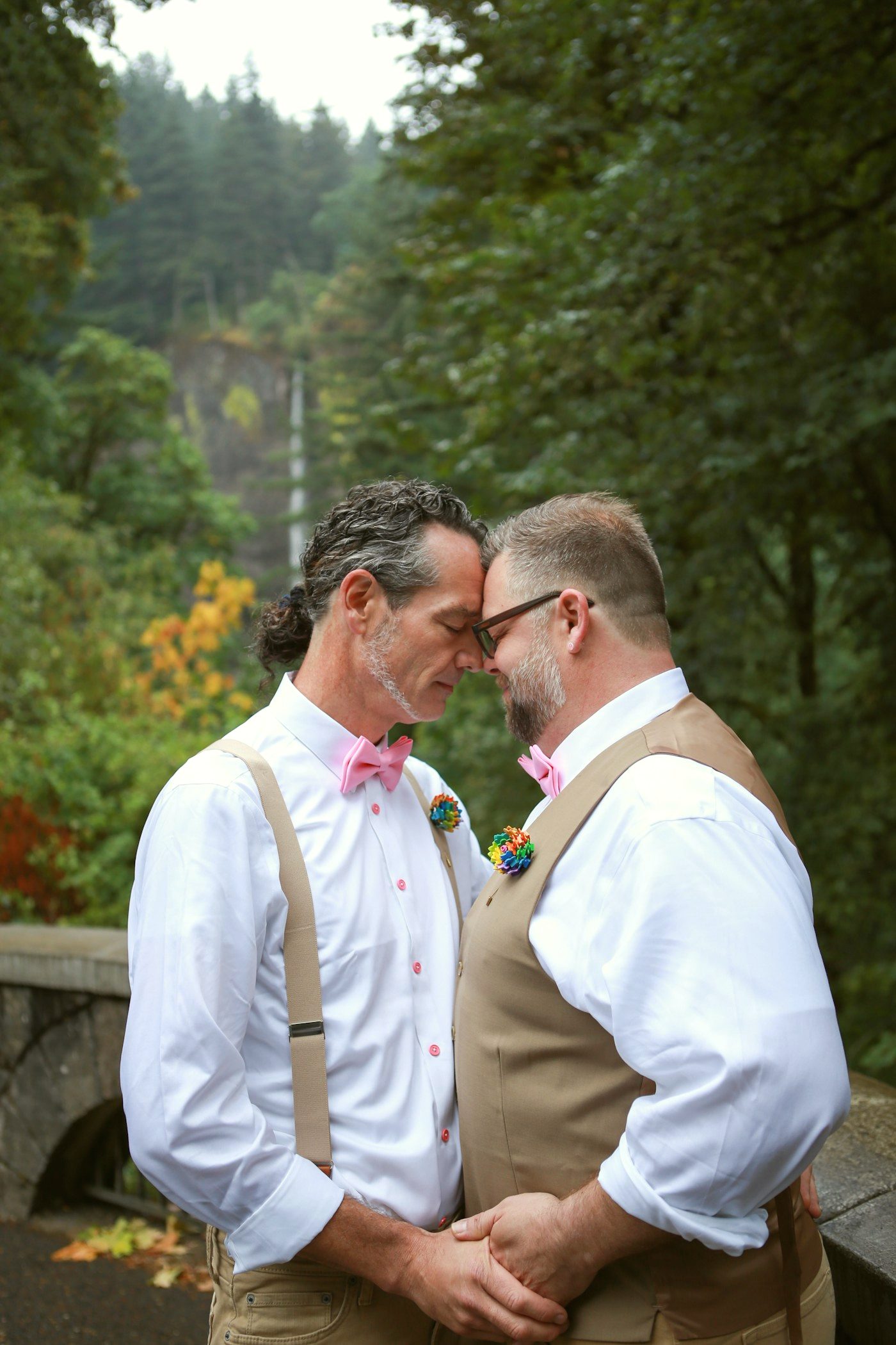 Two grooms with pink bow ties and rainbow boutonnieres, foreheads together